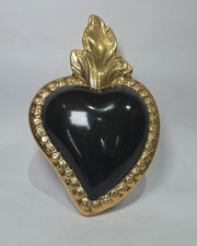 Scultura a forma di Cuore Sacro in ceramica nera lucida, incorniciata da un elegante bordo dorato con decorazioni a rilievo e completata da una fiamma superiore in oro. Questo oggetto di design artigianale con base d'appoggio è un complemento d'arredo raffinato, ideale per arricchire ambienti moderni con un tocco di arte sacra contemporanea.