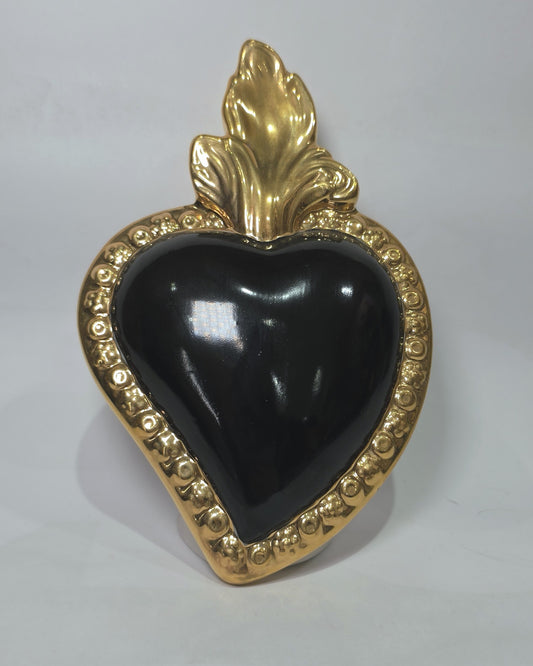 Scultura a forma di Cuore Sacro in ceramica nera lucida, incorniciata da un elegante bordo dorato con decorazioni a rilievo e completata da una fiamma superiore in oro. Questo oggetto di design artigianale con base d'appoggio è un complemento d'arredo raffinato, ideale per arricchire ambienti moderni con un tocco di arte sacra contemporanea.