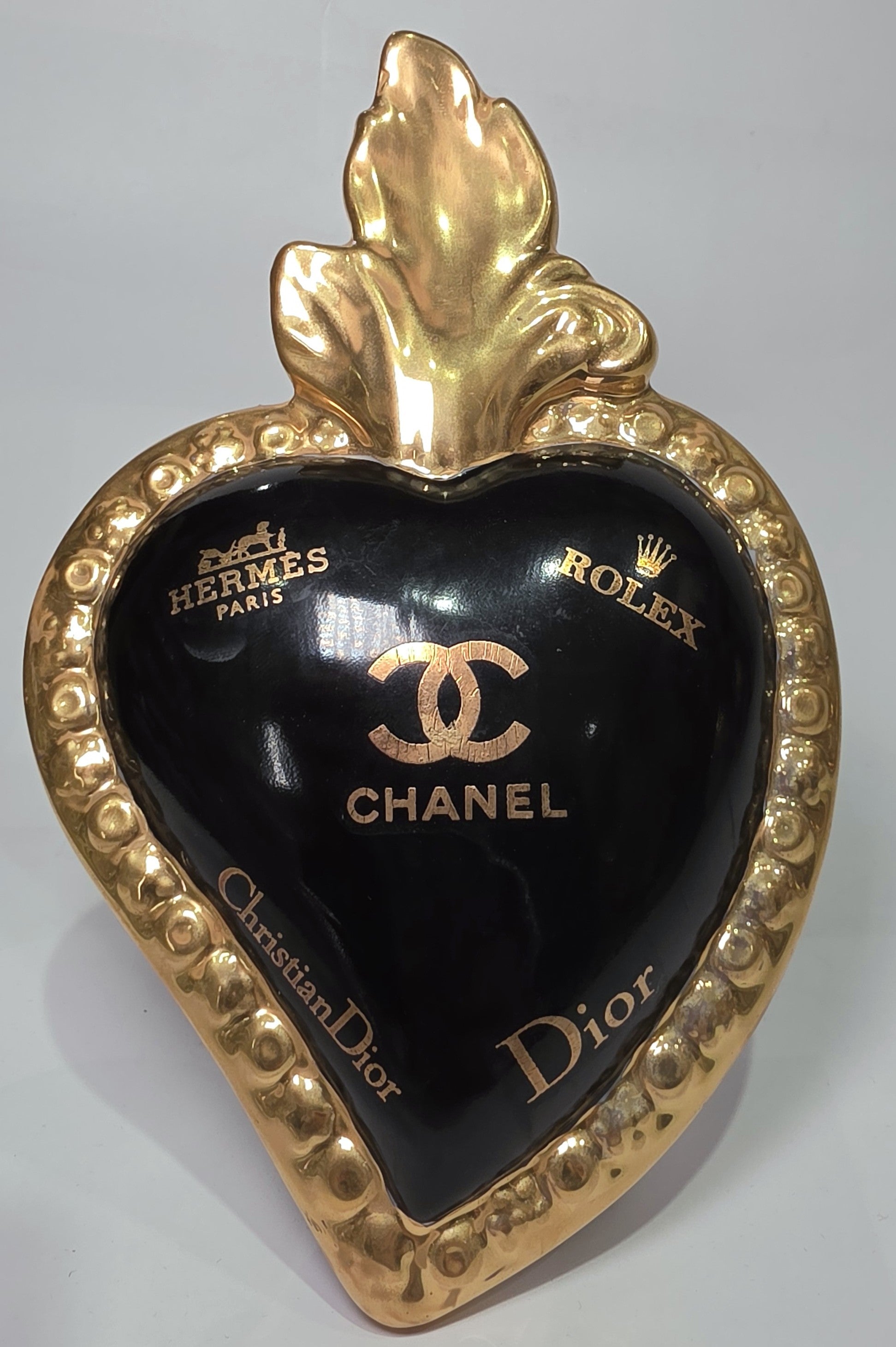 Scultura a forma di Cuore Sacro in ceramica nera lucida, impreziosita da una ricca cornice dorata lavorata a sbalzo e decorata con loghi fashion iconici in oro. Questo esclusivo oggetto d'arredo in stile pop art presenta una fiamma superiore dorata e una base d'appoggio integrata, perfetto per aggiungere un dettaglio di lusso moderno agli ambienti interni.