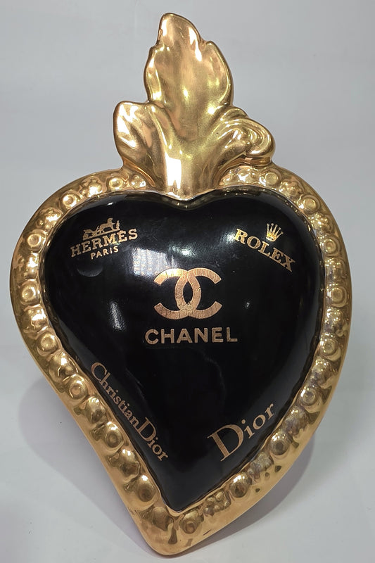 Scultura a forma di Cuore Sacro in ceramica nera lucida, impreziosita da una ricca cornice dorata lavorata a sbalzo e decorata con loghi fashion iconici in oro. Questo esclusivo oggetto d'arredo in stile pop art presenta una fiamma superiore dorata e una base d'appoggio integrata, perfetto per aggiungere un dettaglio di lusso moderno agli ambienti interni.