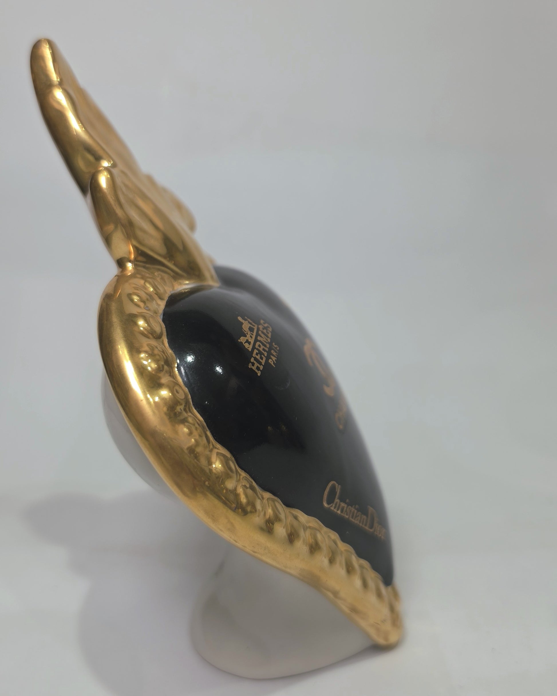 Scultura a forma di Cuore Sacro in ceramica nera lucida, impreziosita da una ricca cornice dorata lavorata a sbalzo e decorata con loghi fashion iconici in oro. Questo esclusivo oggetto d'arredo in stile pop art presenta una fiamma superiore dorata e una base d'appoggio integrata, perfetto per aggiungere un dettaglio di lusso moderno agli ambienti interni.