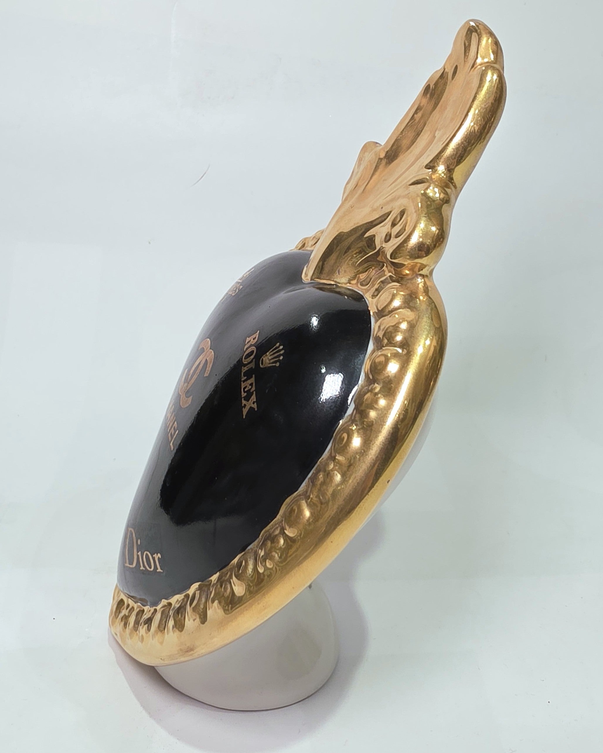 Scultura a forma di Cuore Sacro in ceramica nera lucida, impreziosita da una ricca cornice dorata lavorata a sbalzo e decorata con loghi fashion iconici in oro. Questo esclusivo oggetto d'arredo in stile pop art presenta una fiamma superiore dorata e una base d'appoggio integrata, perfetto per aggiungere un dettaglio di lusso moderno agli ambienti interni.