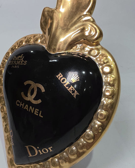 Scultura a forma di Cuore Sacro in ceramica nera lucida, impreziosita da una ricca cornice dorata lavorata a sbalzo e decorata con loghi fashion iconici in oro. Questo esclusivo oggetto d'arredo in stile pop art presenta una fiamma superiore dorata e una base d'appoggio integrata, perfetto per aggiungere un dettaglio di lusso moderno agli ambienti interni.