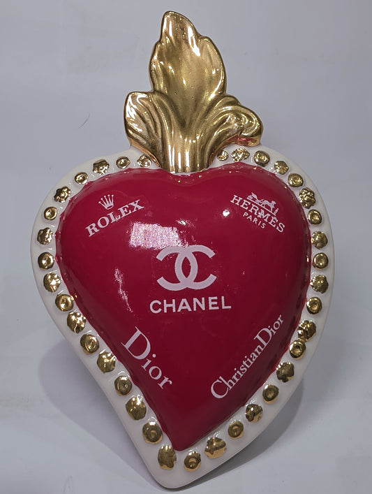 Scultura decorativa a forma di Cuore Sacro in ceramica rossa lucida, arricchita da loghi fashion bianchi e una fiamma superiore dorata. Il design è completato da una cornice bianca perlinata con borchie oro, rendendo questo oggetto un complemento d'arredo pop art unico e di tendenza per la casa.
