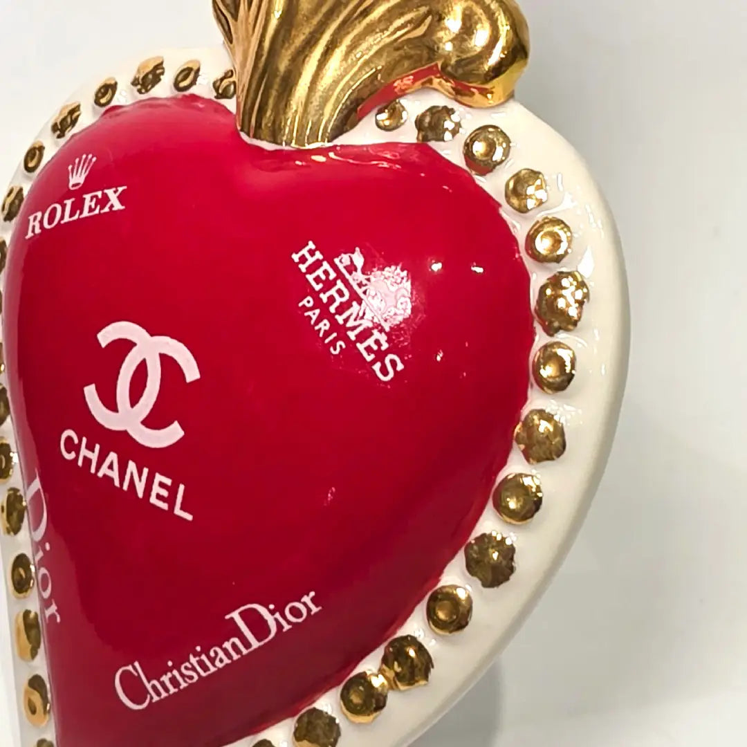 Scultura decorativa a forma di Cuore Sacro in ceramica rossa lucida, arricchita da loghi fashion bianchi e una fiamma superiore dorata. Il design è completato da una cornice bianca perlinata con borchie oro, rendendo questo oggetto un complemento d'arredo pop art unico e di tendenza per la casa.