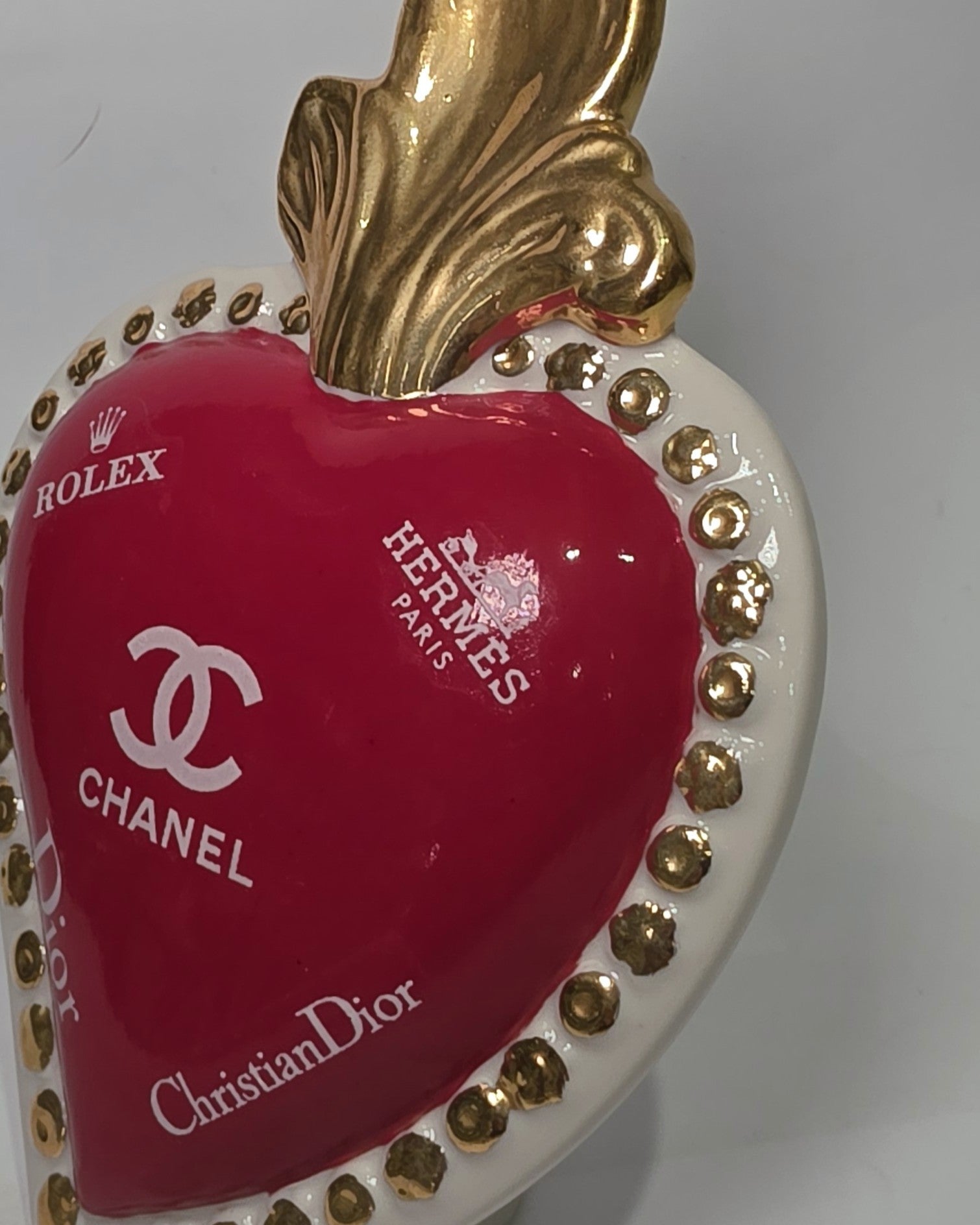 Scultura decorativa a forma di Cuore Sacro in ceramica rossa lucida, arricchita da loghi fashion bianchi e una fiamma superiore dorata. Il design è completato da una cornice bianca perlinata con borchie oro, rendendo questo oggetto un complemento d'arredo pop art unico e di tendenza per la casa.