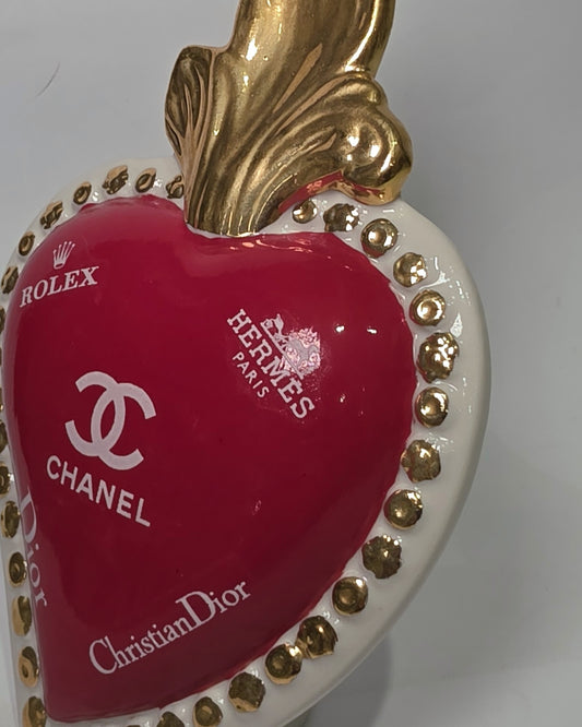 Scultura decorativa a forma di Cuore Sacro in ceramica rossa lucida, arricchita da loghi fashion bianchi e una fiamma superiore dorata. Il design è completato da una cornice bianca perlinata con borchie oro, rendendo questo oggetto un complemento d'arredo pop art unico e di tendenza per la casa.