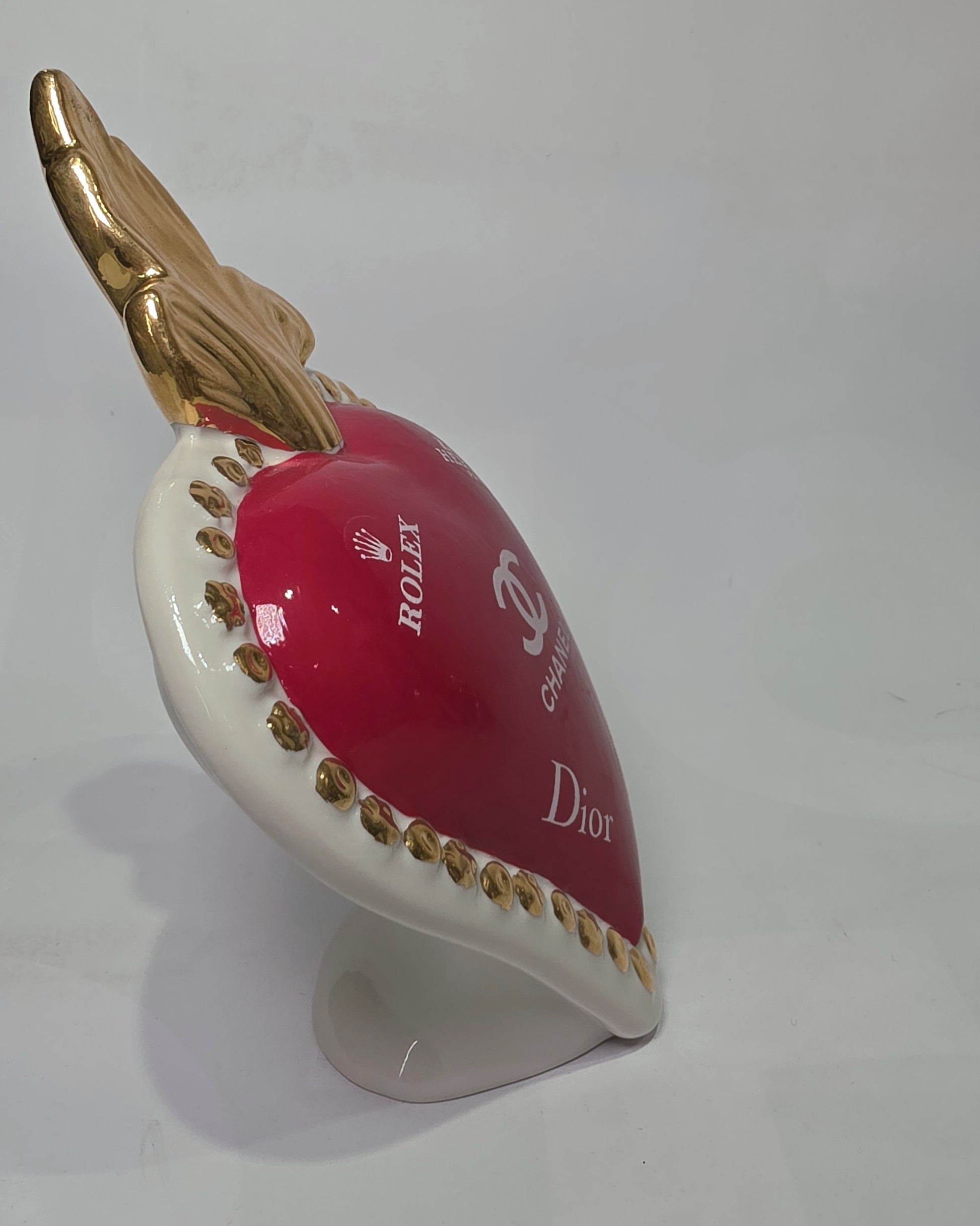 Scultura decorativa a forma di Cuore Sacro in ceramica rossa lucida, arricchita da loghi fashion bianchi e una fiamma superiore dorata. Il design è completato da una cornice bianca perlinata con borchie oro, rendendo questo oggetto un complemento d'arredo pop art unico e di tendenza per la casa.