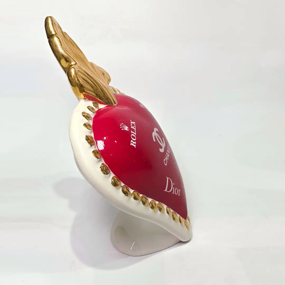 Scultura decorativa a forma di Cuore Sacro in ceramica rossa lucida, arricchita da loghi fashion bianchi e una fiamma superiore dorata. Il design è completato da una cornice bianca perlinata con borchie oro, rendendo questo oggetto un complemento d'arredo pop art unico e di tendenza per la casa.