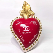 Scultura decorativa a forma di Cuore Sacro in ceramica rossa lucida, arricchita da loghi fashion bianchi e una fiamma superiore dorata. Il design è completato da una cornice bianca perlinata con borchie oro, rendendo questo oggetto un complemento d'arredo pop art unico e di tendenza per la casa.