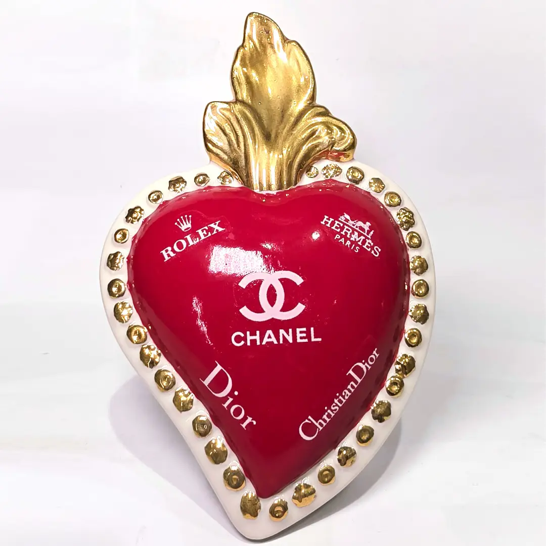 Scultura decorativa a forma di Cuore Sacro in ceramica rossa lucida, arricchita da loghi fashion bianchi e una fiamma superiore dorata. Il design è completato da una cornice bianca perlinata con borchie oro, rendendo questo oggetto un complemento d'arredo pop art unico e di tendenza per la casa.