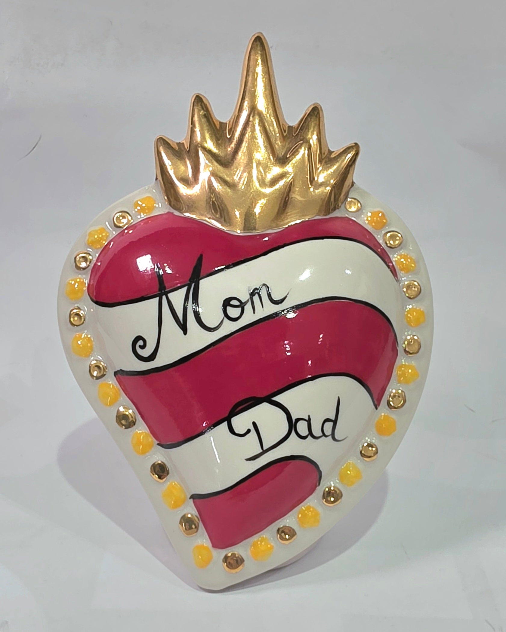 Scultura decorativa a forma di Cuore Sacro in ceramica rossa e bianca lucida, caratterizzata da un design a strisce orizzontali con le scritte "Mom" e "Dad" in corsivo nero. L'oggetto è completato da una corona superiore dorata e una cornice perlinata con dettagli gialli e borchie oro, rappresentando un complemento d'arredo affettivo e originale per la casa.