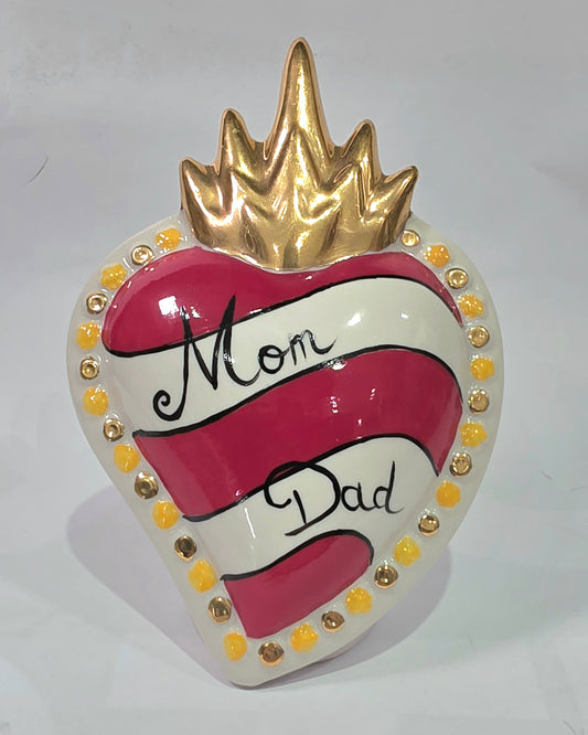 Scultura decorativa a forma di Cuore Sacro in ceramica rossa e bianca lucida, caratterizzata da un design a strisce orizzontali con le scritte "Mom" e "Dad" in corsivo nero. L'oggetto è completato da una corona superiore dorata e una cornice perlinata con dettagli gialli e borchie oro, rappresentando un complemento d'arredo affettivo e originale per la casa.