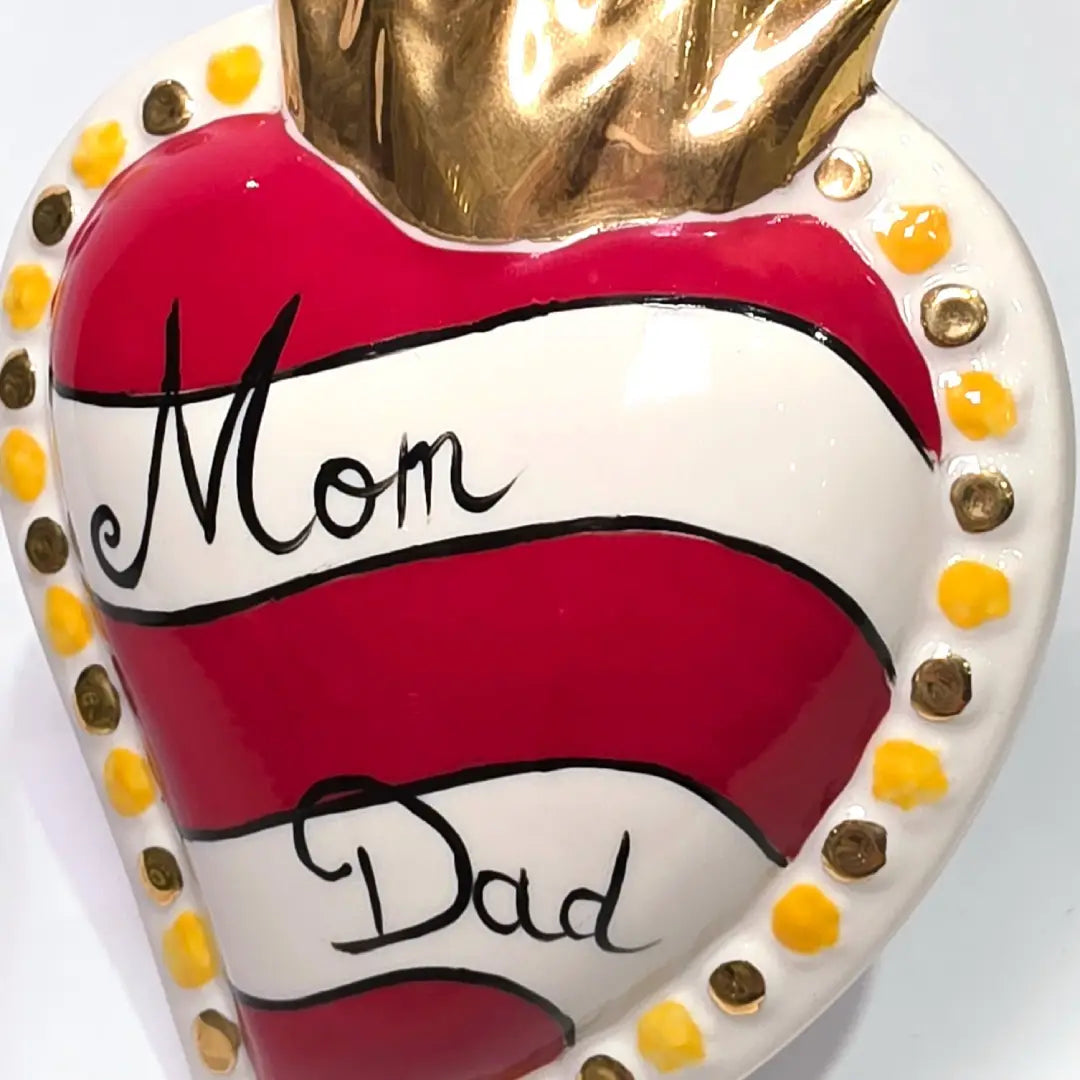 Scultura decorativa a forma di Cuore Sacro in ceramica rossa e bianca lucida, caratterizzata da un design a strisce orizzontali con le scritte "Mom" e "Dad" in corsivo nero. L'oggetto è completato da una corona superiore dorata e una cornice perlinata con dettagli gialli e borchie oro, rappresentando un complemento d'arredo affettivo e originale per la casa.