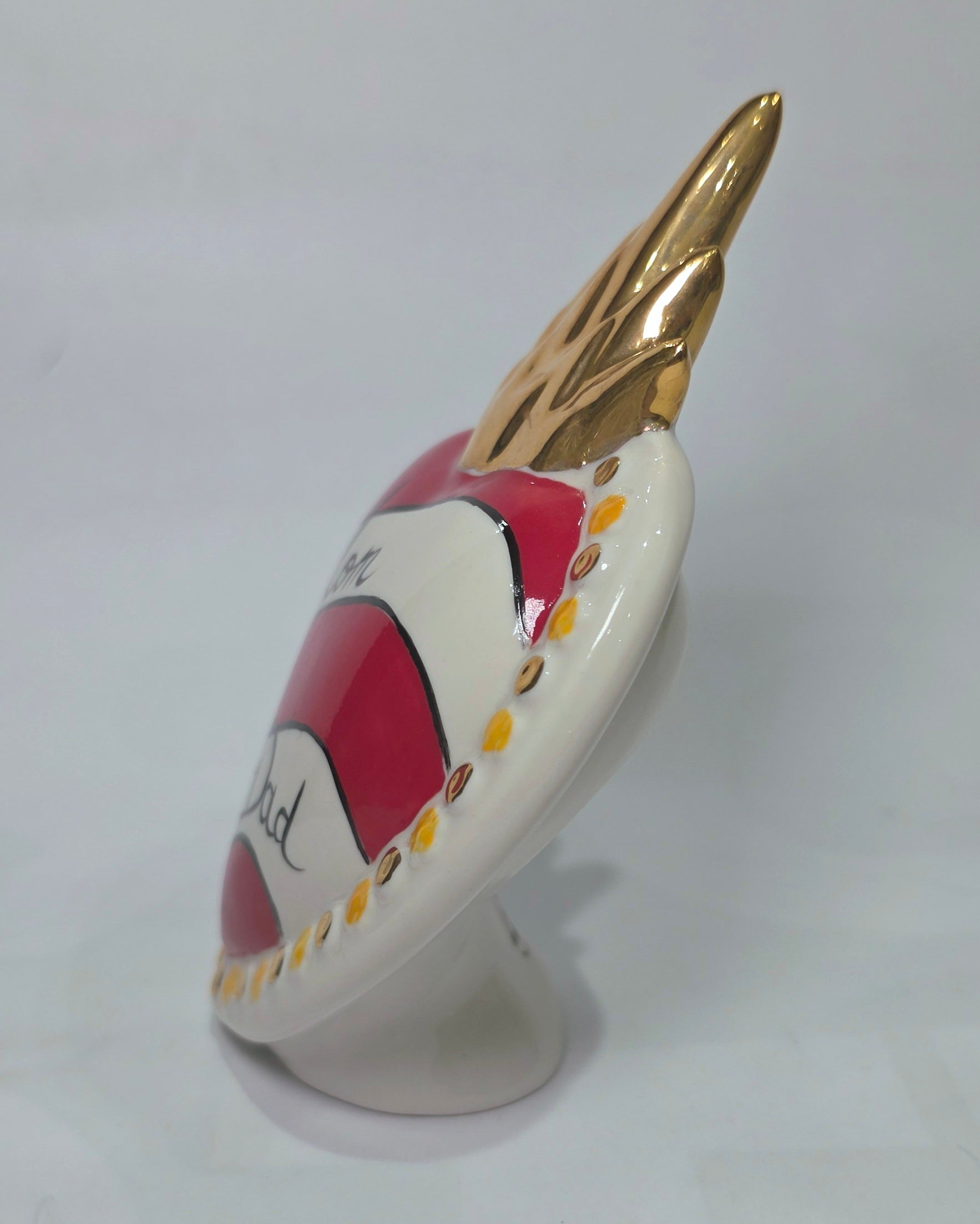 Scultura decorativa a forma di Cuore Sacro in ceramica rossa e bianca lucida, caratterizzata da un design a strisce orizzontali con le scritte "Mom" e "Dad" in corsivo nero. L'oggetto è completato da una corona superiore dorata e una cornice perlinata con dettagli gialli e borchie oro, rappresentando un complemento d'arredo affettivo e originale per la casa.