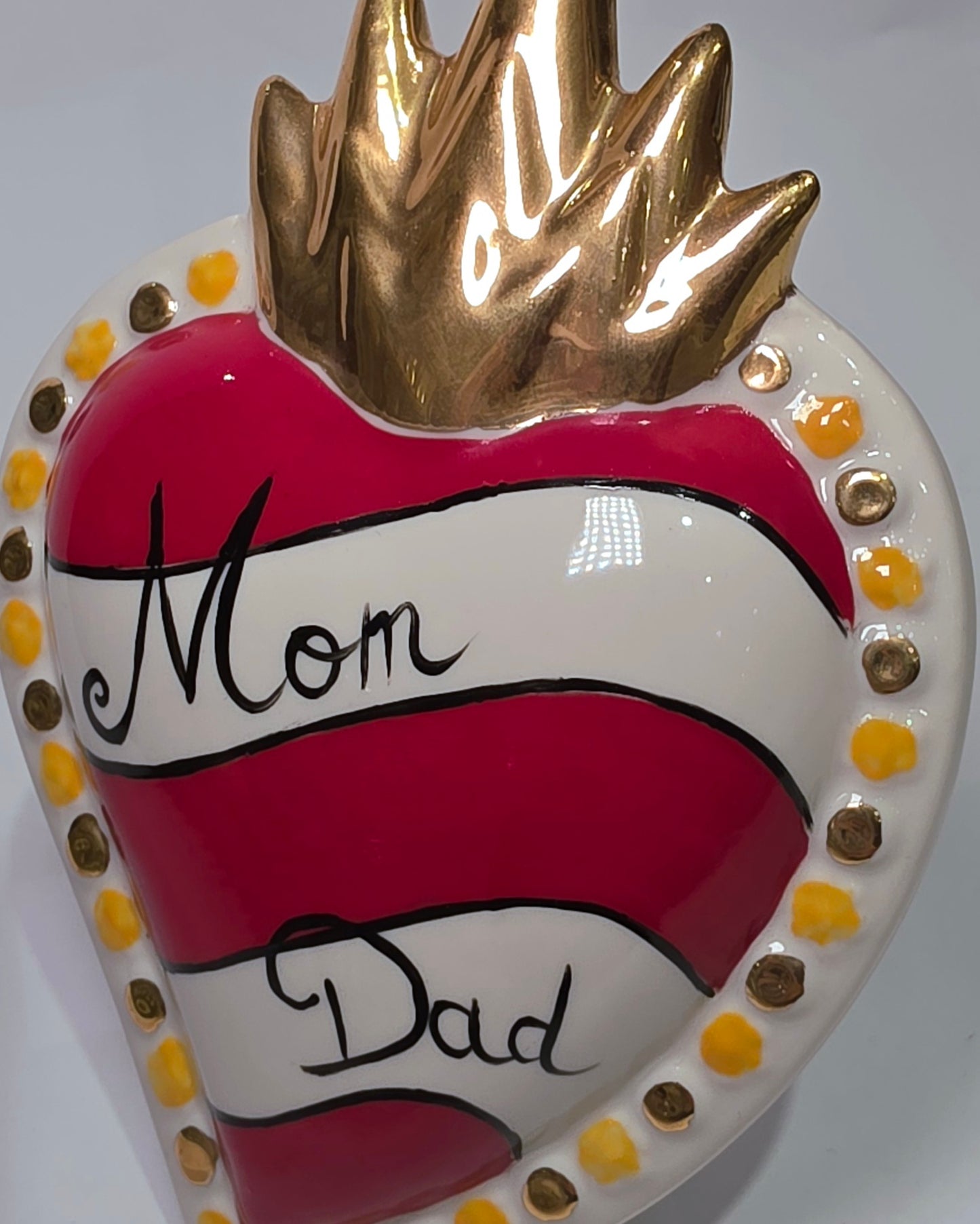 Scultura decorativa a forma di Cuore Sacro in ceramica rossa e bianca lucida, caratterizzata da un design a strisce orizzontali con le scritte "Mom" e "Dad" in corsivo nero. L'oggetto è completato da una corona superiore dorata e una cornice perlinata con dettagli gialli e borchie oro, rappresentando un complemento d'arredo affettivo e originale per la casa.