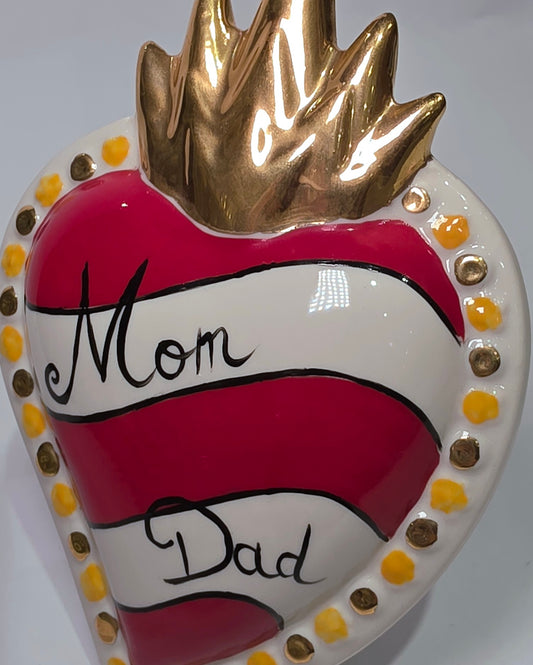 Scultura decorativa a forma di Cuore Sacro in ceramica rossa e bianca lucida, caratterizzata da un design a strisce orizzontali con le scritte "Mom" e "Dad" in corsivo nero. L'oggetto è completato da una corona superiore dorata e una cornice perlinata con dettagli gialli e borchie oro, rappresentando un complemento d'arredo affettivo e originale per la casa.