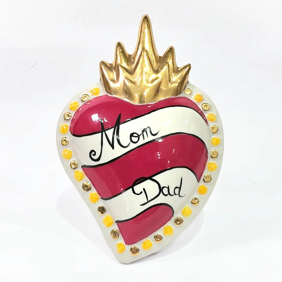 Scultura decorativa a forma di Cuore Sacro in ceramica rossa e bianca lucida, caratterizzata da un design a strisce orizzontali con le scritte "Mom" e "Dad" in corsivo nero. L'oggetto è completato da una corona superiore dorata e una cornice perlinata con dettagli gialli e borchie oro, rappresentando un complemento d'arredo affettivo e originale per la casa.