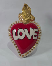Scultura decorativa a forma di Cuore Sacro in ceramica rossa lucida, caratterizzata dalla scritta "LOVE" in stile pop art bianco e nero al centro del quadrante. L'opera è rifinita con una fiamma superiore dorata e una cornice bianca decorata con borchie oro, perfetta come complemento d'arredo moderno o idea regalo romantica.