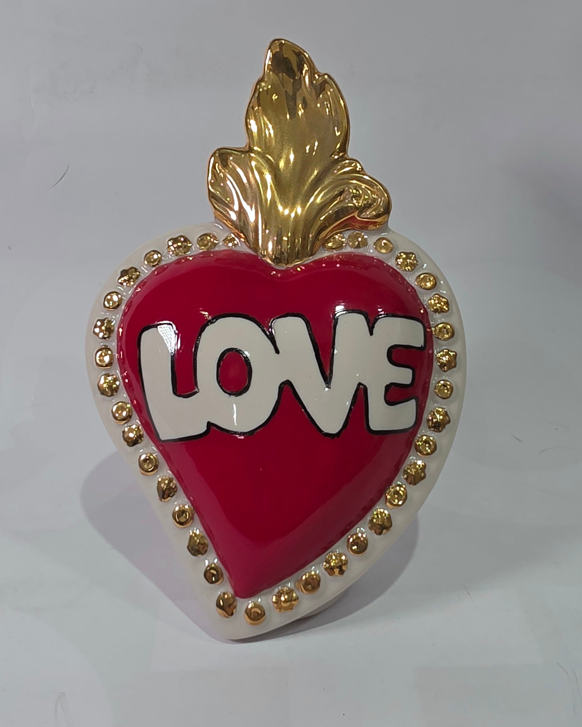 Scultura decorativa a forma di Cuore Sacro in ceramica rossa lucida, caratterizzata dalla scritta "LOVE" in stile pop art bianco e nero al centro del quadrante. L'opera è rifinita con una fiamma superiore dorata e una cornice bianca decorata con borchie oro, perfetta come complemento d'arredo moderno o idea regalo romantica.