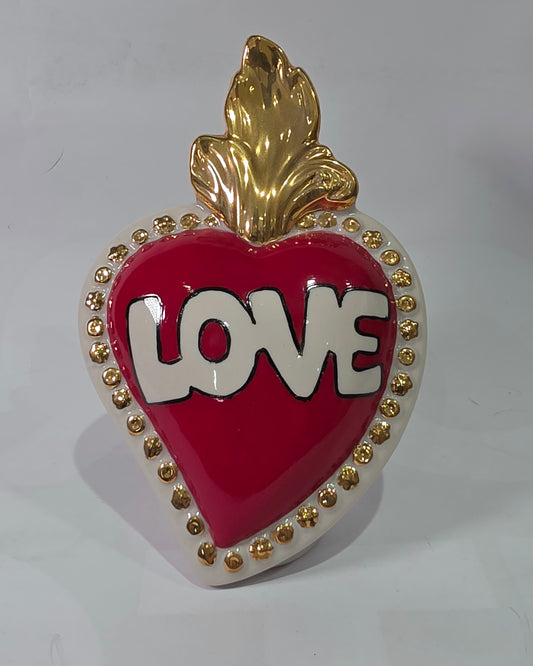 Scultura decorativa a forma di Cuore Sacro in ceramica rossa lucida, caratterizzata dalla scritta "LOVE" in stile pop art bianco e nero al centro del quadrante. L'opera è rifinita con una fiamma superiore dorata e una cornice bianca decorata con borchie oro, perfetta come complemento d'arredo moderno o idea regalo romantica.