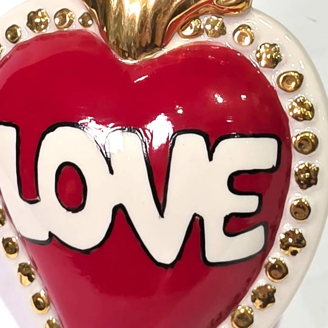 Scultura decorativa a forma di Cuore Sacro in ceramica rossa lucida, caratterizzata dalla scritta "LOVE" in stile pop art bianco e nero al centro del quadrante. L'opera è rifinita con una fiamma superiore dorata e una cornice bianca decorata con borchie oro, perfetta come complemento d'arredo moderno o idea regalo romantica.