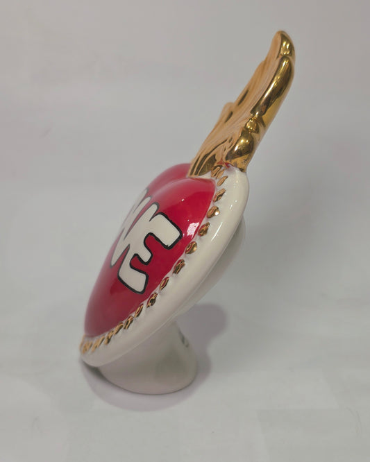 Scultura decorativa a forma di Cuore Sacro in ceramica rossa lucida, caratterizzata dalla scritta "LOVE" in stile pop art bianco e nero al centro del quadrante. L'opera è rifinita con una fiamma superiore dorata e una cornice bianca decorata con borchie oro, perfetta come complemento d'arredo moderno o idea regalo romantica.
