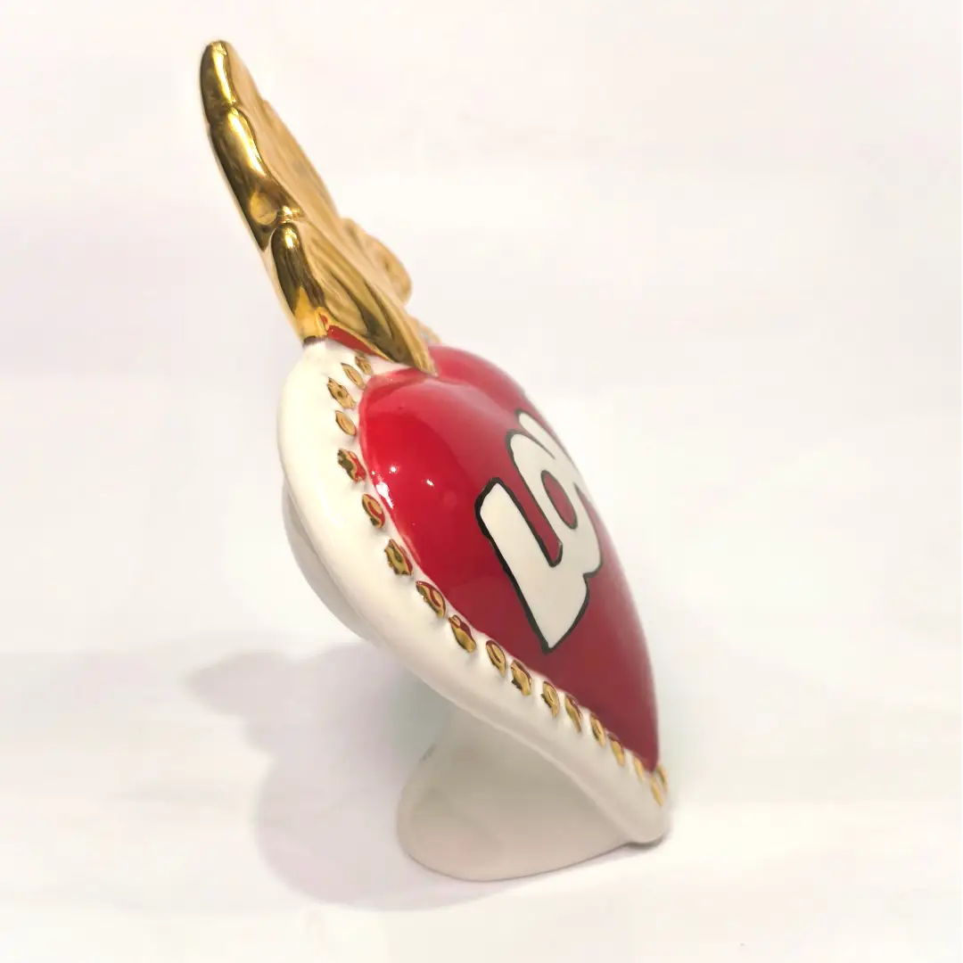 Scultura decorativa a forma di Cuore Sacro in ceramica rossa lucida, caratterizzata dalla scritta "LOVE" in stile pop art bianco e nero al centro del quadrante. L'opera è rifinita con una fiamma superiore dorata e una cornice bianca decorata con borchie oro, perfetta come complemento d'arredo moderno o idea regalo romantica.