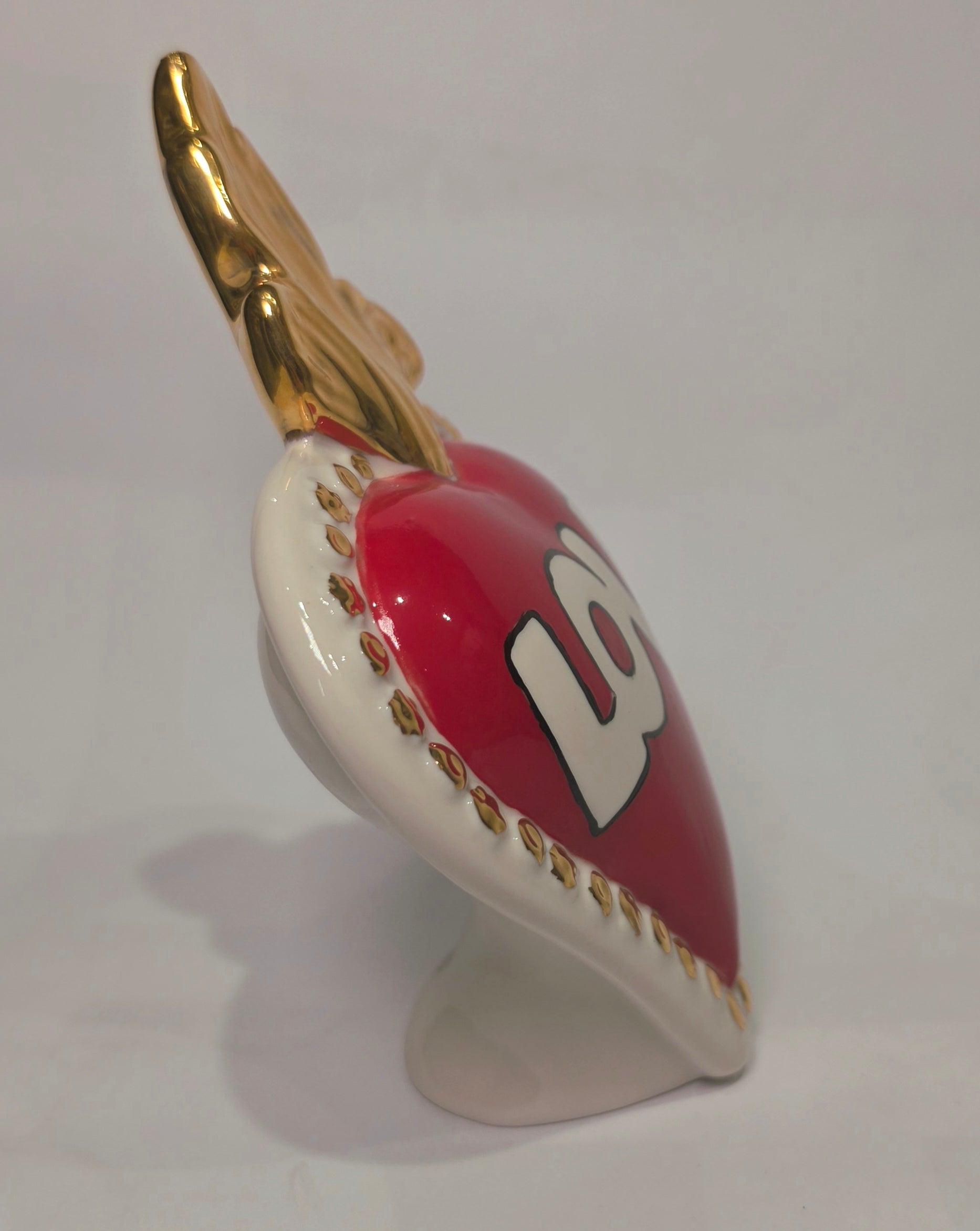Scultura decorativa a forma di Cuore Sacro in ceramica rossa lucida, caratterizzata dalla scritta "LOVE" in stile pop art bianco e nero al centro del quadrante. L'opera è rifinita con una fiamma superiore dorata e una cornice bianca decorata con borchie oro, perfetta come complemento d'arredo moderno o idea regalo romantica.
