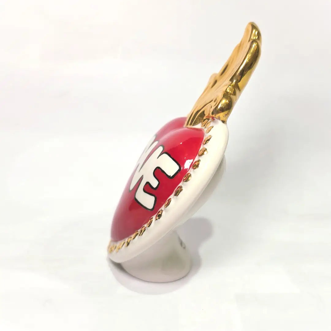 Scultura decorativa a forma di Cuore Sacro in ceramica rossa lucida, caratterizzata dalla scritta "LOVE" in stile pop art bianco e nero al centro del quadrante. L'opera è rifinita con una fiamma superiore dorata e una cornice bianca decorata con borchie oro, perfetta come complemento d'arredo moderno o idea regalo romantica.
