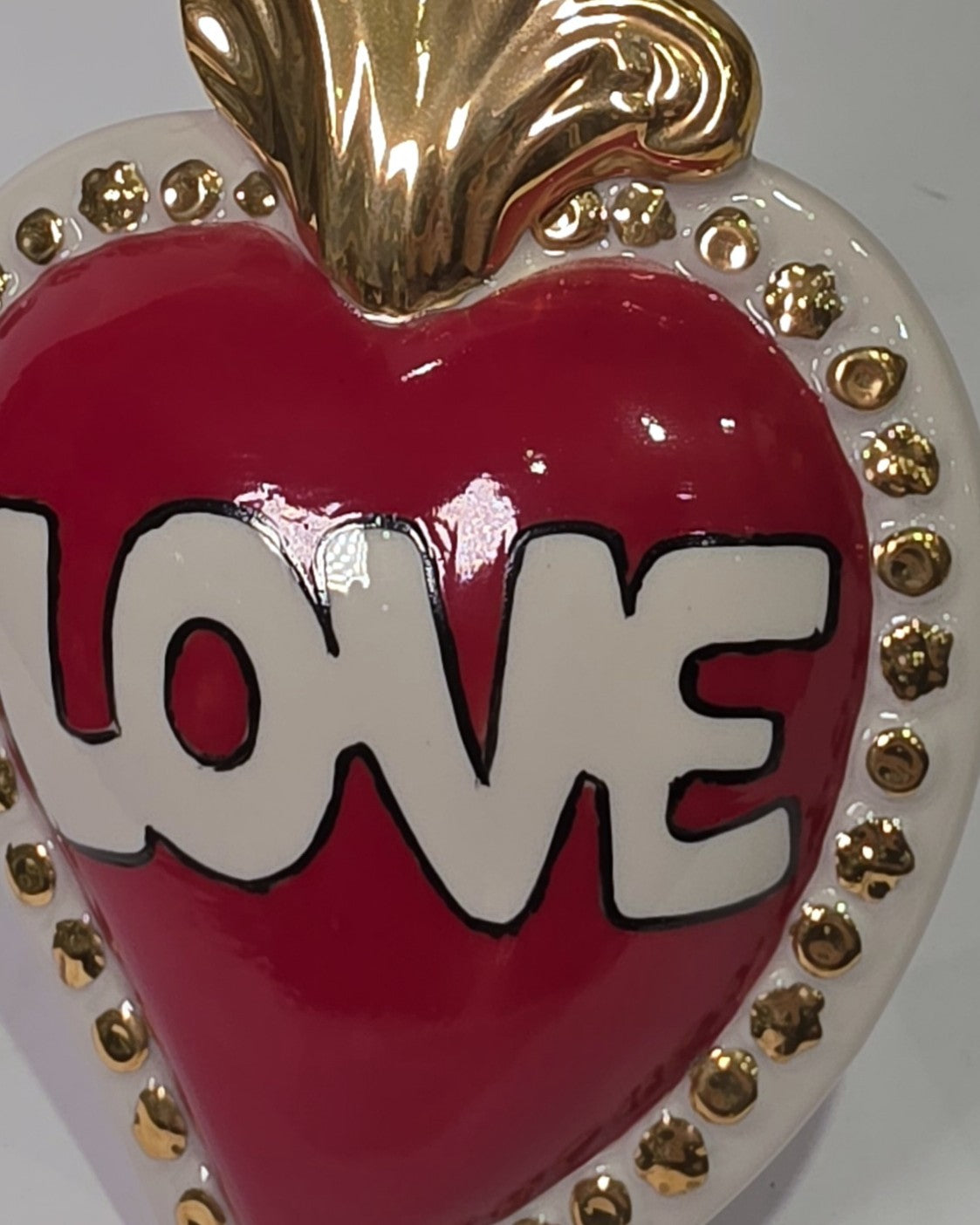 Scultura decorativa a forma di Cuore Sacro in ceramica rossa lucida, caratterizzata dalla scritta "LOVE" in stile pop art bianco e nero al centro del quadrante. L'opera è rifinita con una fiamma superiore dorata e una cornice bianca decorata con borchie oro, perfetta come complemento d'arredo moderno o idea regalo romantica.