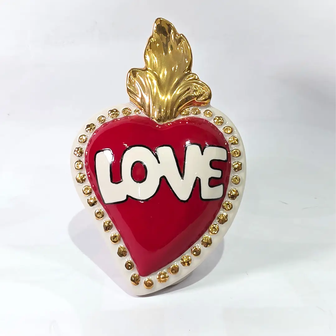 Scultura decorativa a forma di Cuore Sacro in ceramica rossa lucida, caratterizzata dalla scritta "LOVE" in stile pop art bianco e nero al centro del quadrante. L'opera è rifinita con una fiamma superiore dorata e una cornice bianca decorata con borchie oro, perfetta come complemento d'arredo moderno o idea regalo romantica.