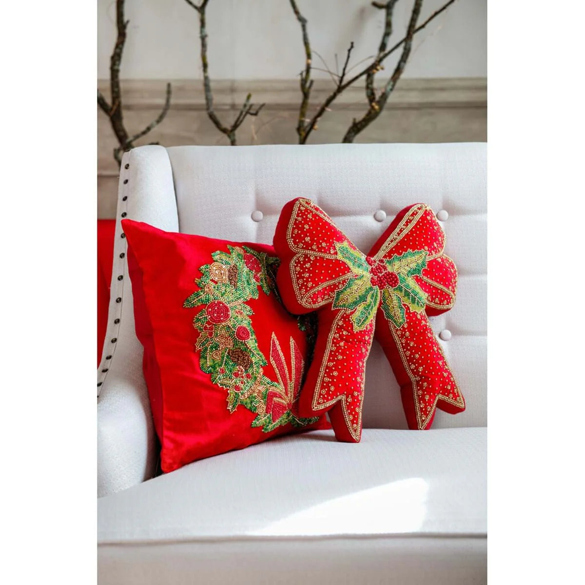 Set di cuscini natalizi rossi su una poltrona bianca classica. Include un cuscino quadrato con ghirlanda ricamata e un cuscino a forma di fiocco con agrifoglio. Atmosfera festiva elegante e accogliente.