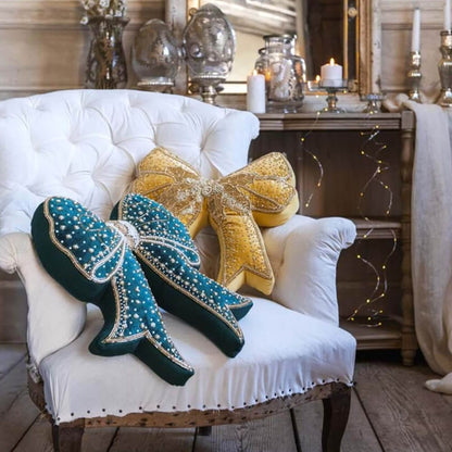 Coppia di cuscini a fiocco Blanc Mariclò in velluto verde e oro adagiati su una poltrona capitonné bianca. Atmosfera elegante con candele e arredi shabby chic sullo sfondo.
