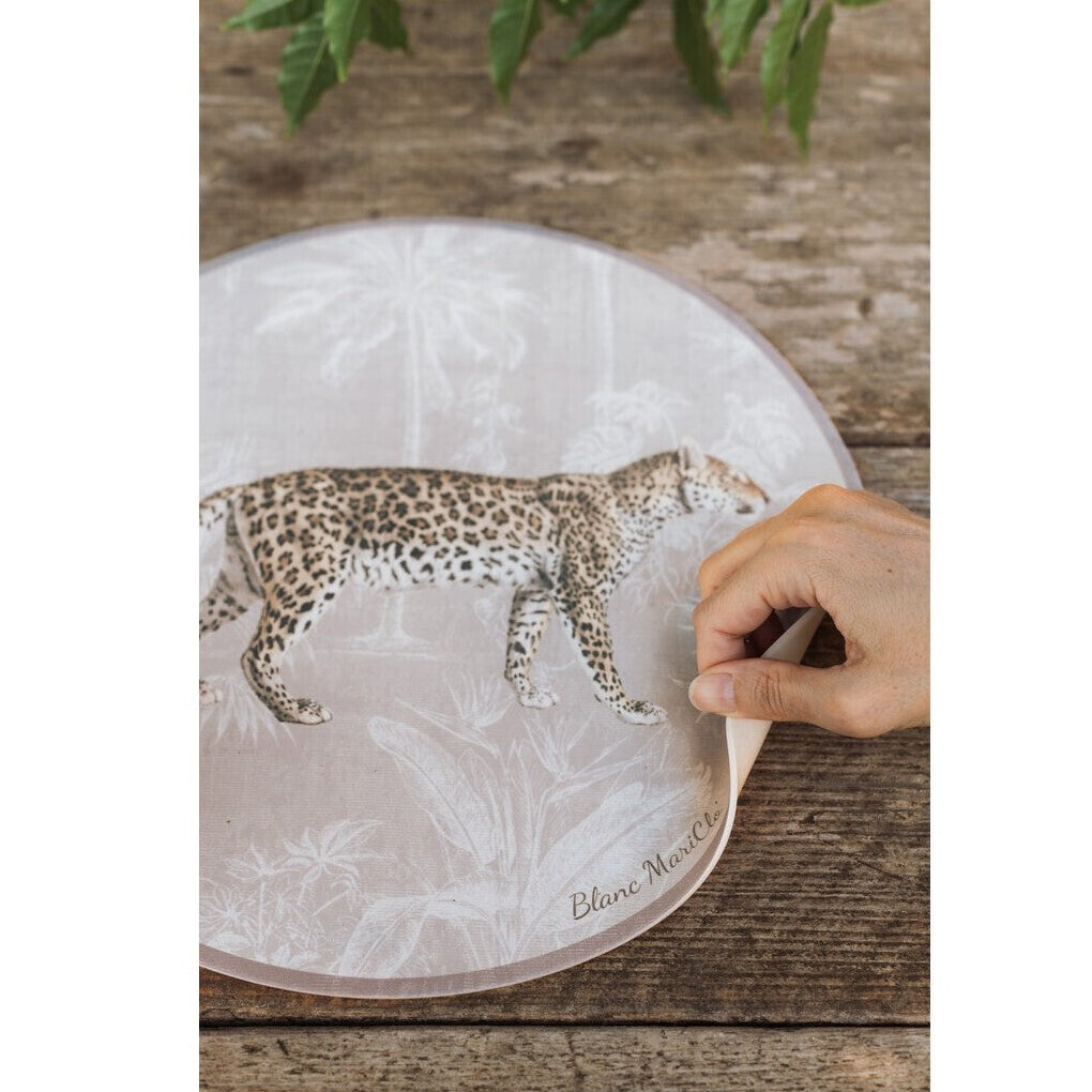 Tovaglietta americana rotonda in vinile della collezione "Leopard" di Blanc Mariclo' caratterizzata dall'illustrazione realistica di un leopardo maculato in posizione di profilo. Lo sfondo beige presenta un delicato motivo esotico con palme e piante tropicali stilizzate in bianco, ideale per apparecchiare una tavola moderna e grintosa dal tocco jungle chic.