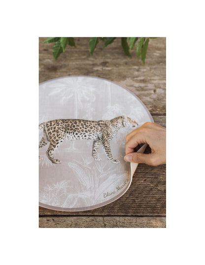 Tovaglietta americana rotonda in vinile della collezione "Leopard" di Blanc Mariclo' caratterizzata dall'illustrazione realistica di un leopardo maculato in posizione di profilo. Lo sfondo beige presenta un delicato motivo esotico con palme e piante tropicali stilizzate in bianco, ideale per apparecchiare una tavola moderna e grintosa dal tocco jungle chic.