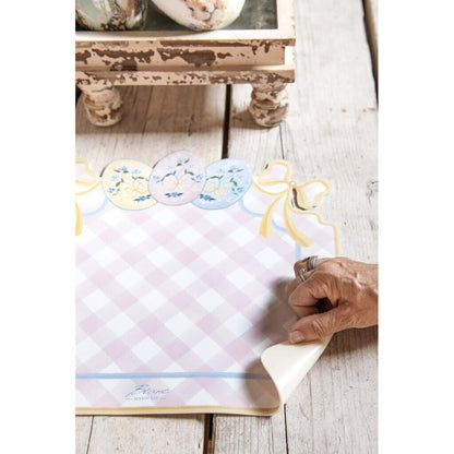 Tovaglietta americana in vinile della collezione "Pretty Easter" firmata Blanc Mariclo' con un motivo a scacchi bianchi e rosa perfetto per la colazione o il pranzo pasquale. La tovaglietta sagomata è decorata sul bordo superiore con fiocchi gialli e tre uova di Pasqua colorate nei toni pastello con dettagli floreali azzurri.