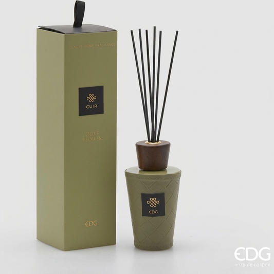 Il Diffusore per Ambienti Cuir Olive Flower EDG è un set di profumazione per la casa composto da una bottiglia in vetro sfaccettato verde scuro, impreziosita da un rivestimento protettivo marrone con cuciture a contrasto e un'etichetta dorata. La confezione cilindrica coordinata con dettagli oro racchiude i bastoncini diffusori neri, rendendo questa fragranza un elemento di home decor elegante e perfetto come idea regalo esclusiva.