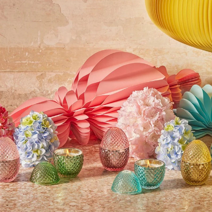 Immagine d'ambientazione per la collezione di candele profumate "L'Ove" firmata EDG Enzo de Gasperi, con eleganti contenitori in vetro colorato lavorato a rilievo a forma di uovo disposti tra decorazioni floreali e festoni di carta colorati. Questa linea di candele decorative di lusso, mostrata sia aperte e accese che con coperchio coordinato, è ideale per profumare gli interni con eleganza e creare un'atmosfera allegra, ricercata e primaverile.
