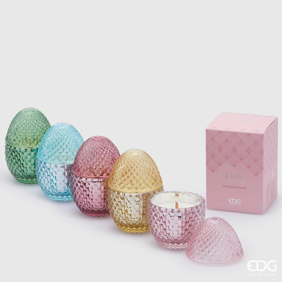 Immagine d'ambientazione per la collezione di candele profumate "L'Ove" firmata EDG Enzo de Gasperi, con eleganti contenitori in vetro colorato lavorato a rilievo a forma di uovo disposti tra decorazioni floreali e festoni di carta colorati. Questa linea di candele decorative di lusso, mostrata sia aperte e accese che con coperchio coordinato, è ideale per profumare gli interni con eleganza e creare un'atmosfera allegra, ricercata e primaverile.