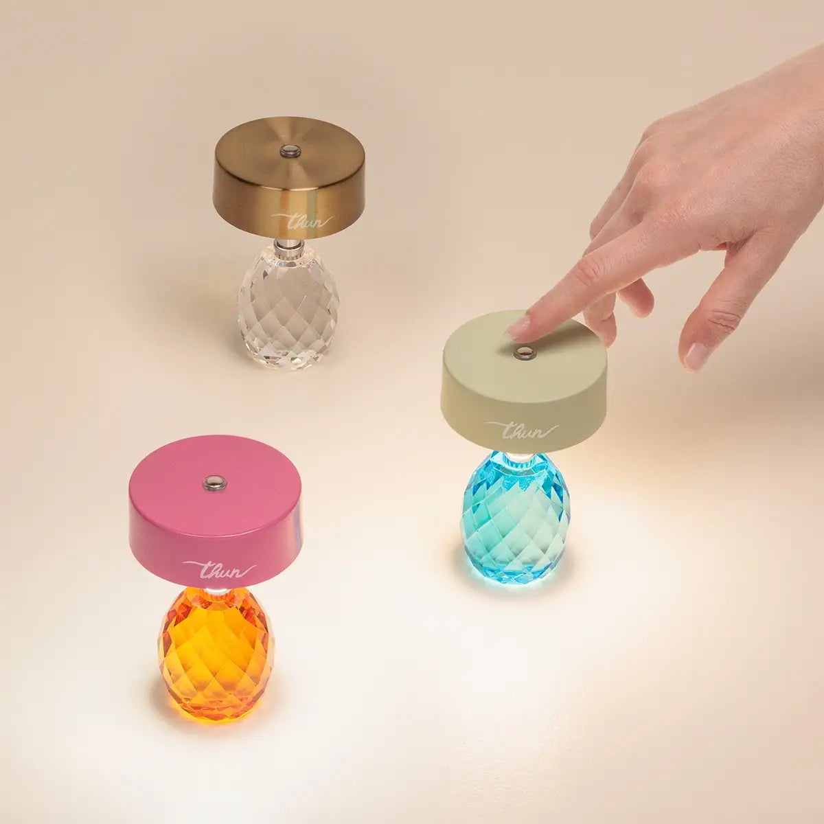 Tre lampade ricaricabile Thun in diverse combinazioni di colore disposte su un piano: una oro con cristallo trasparente, una rosa con cristallo arancione e una verde con cristallo azzurro. Una mano sta toccando il sensore superiore della lampada verde per accenderla.