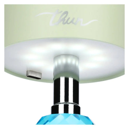 Inquadratura dal basso del paralume verde che mostra il pannello LED circolare acceso, il quale emette una luce calda attraverso il diffusore bianco. Sono visibili anche la porta USB-C, lo stelo cromato e la parte superiore del cristallo azzurro.