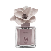 Profumatore d'ambiente Muhà Magnolia: flacone quadrato in vetro trasparente con essenza Ambra Antica e scenografico fiore in betulla color viola grigio.