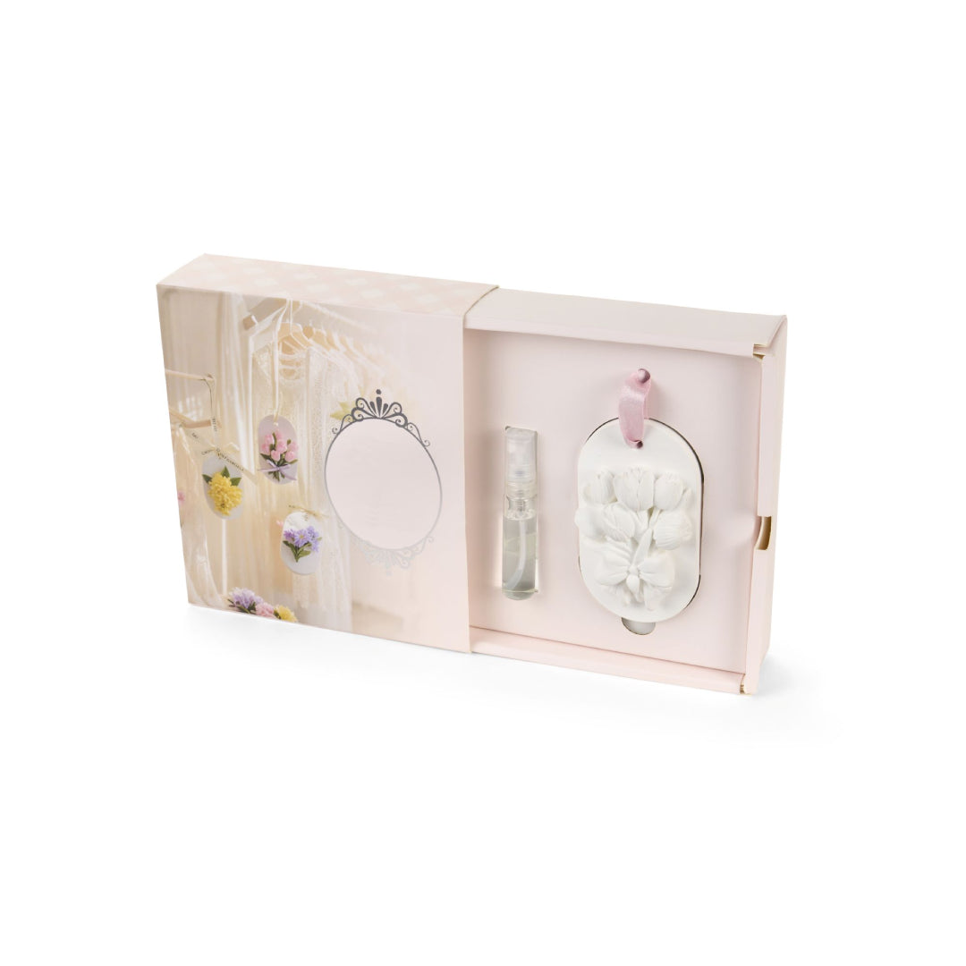 Gessetto profumato da appendere della collezione "Fiorirà" di Nuvole di Stoffa, caratterizzato da una placchetta ovale bianca decorata con un bouquet di tulipani in rilievo e un nastrino rosa. Il set include una boccetta di profumo spray concentrato ed è presentato in un'elegante confezione a cassetto nei toni del rosa pastello, ideale come bomboniera o profumatore per armadi e cassetti.