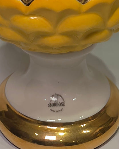 Pigna portafortuna in ceramica bicolore gialla e bianca lucida, impreziosita da una raffinata corona di squame dorate che ne definisce il profilo centrale. Questo elegante oggetto ornamentale artigianale poggia su una base circolare con finiture in oro, rappresentando un complemento d'arredo perfetto per decorazioni d'interni in stile chic e contemporaneo.