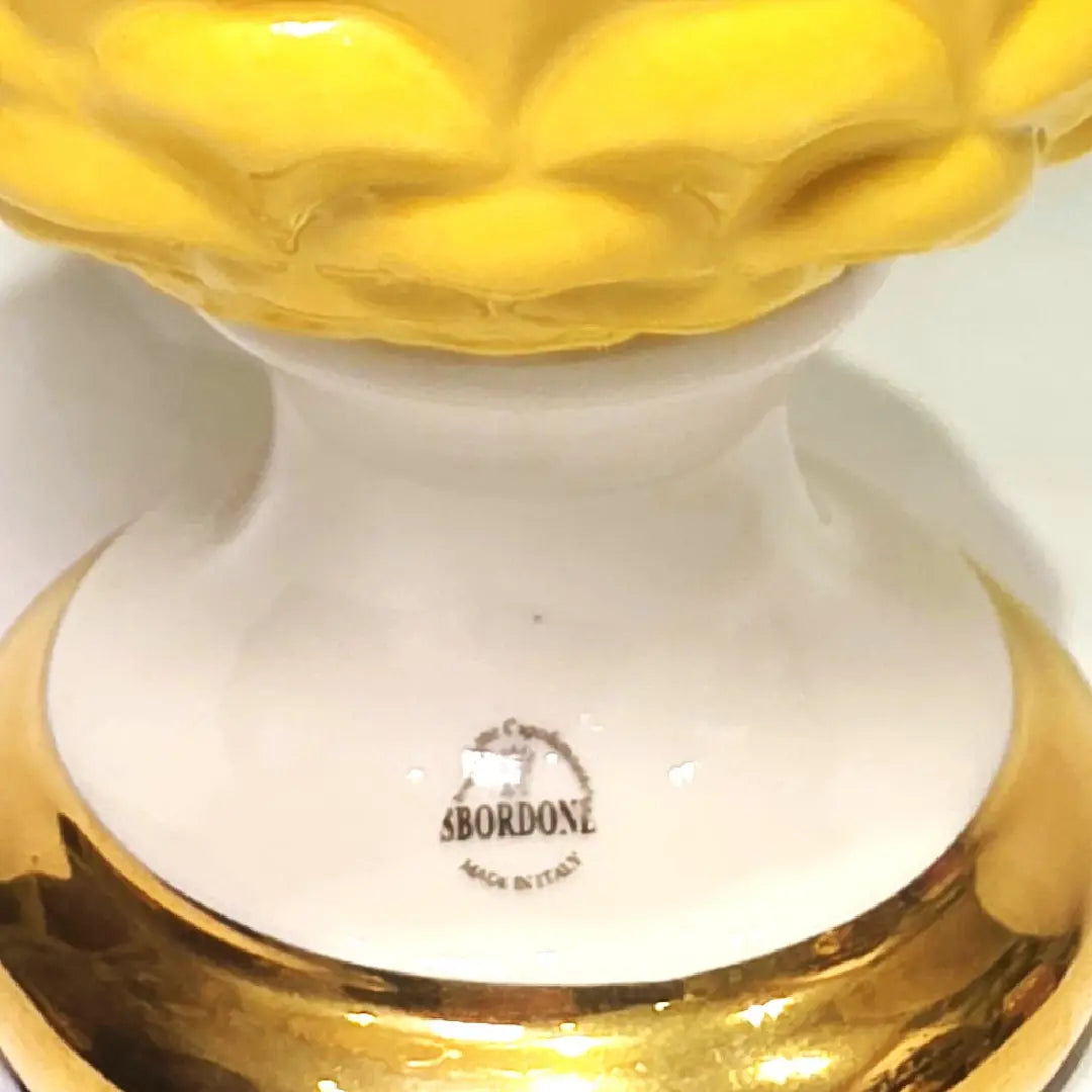 Pigna portafortuna in ceramica bicolore gialla e bianca lucida, impreziosita da una raffinata corona di squame dorate che ne definisce il profilo centrale. Questo elegante oggetto ornamentale artigianale poggia su una base circolare con finiture in oro, rappresentando un complemento d'arredo perfetto per decorazioni d'interni in stile chic e contemporaneo.