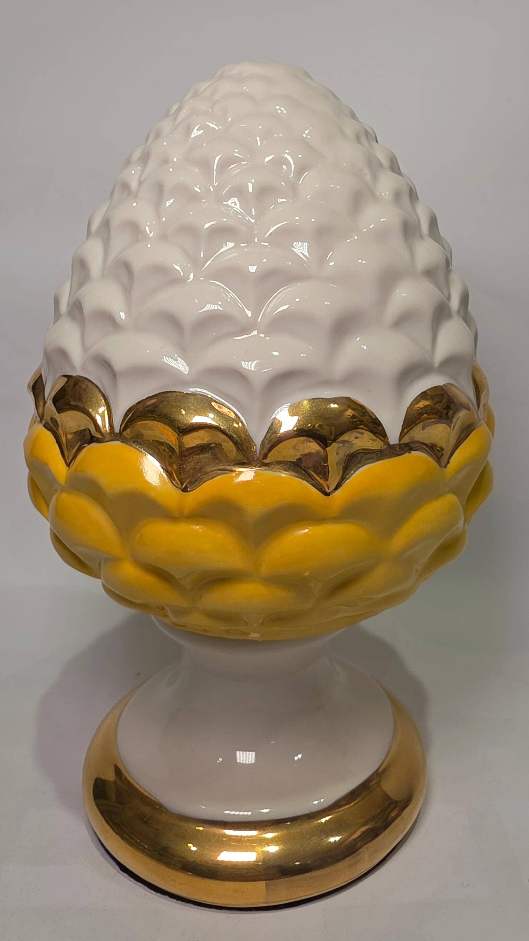Pigna portafortuna in ceramica bicolore gialla e bianca lucida, impreziosita da una raffinata corona di squame dorate che ne definisce il profilo centrale. Questo elegante oggetto ornamentale artigianale poggia su una base circolare con finiture in oro, rappresentando un complemento d'arredo perfetto per decorazioni d'interni in stile chic e contemporaneo.