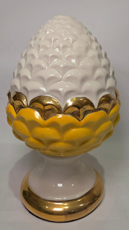 Pigna portafortuna in ceramica bicolore gialla e bianca lucida, impreziosita da una raffinata corona di squame dorate che ne definisce il profilo centrale. Questo elegante oggetto ornamentale artigianale poggia su una base circolare con finiture in oro, rappresentando un complemento d'arredo perfetto per decorazioni d'interni in stile chic e contemporaneo.