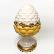 Pigna portafortuna in ceramica bicolore gialla e bianca lucida, impreziosita da una raffinata corona di squame dorate che ne definisce il profilo centrale. Questo elegante oggetto ornamentale artigianale poggia su una base circolare con finiture in oro, rappresentando un complemento d'arredo perfetto per decorazioni d'interni in stile chic e contemporaneo.