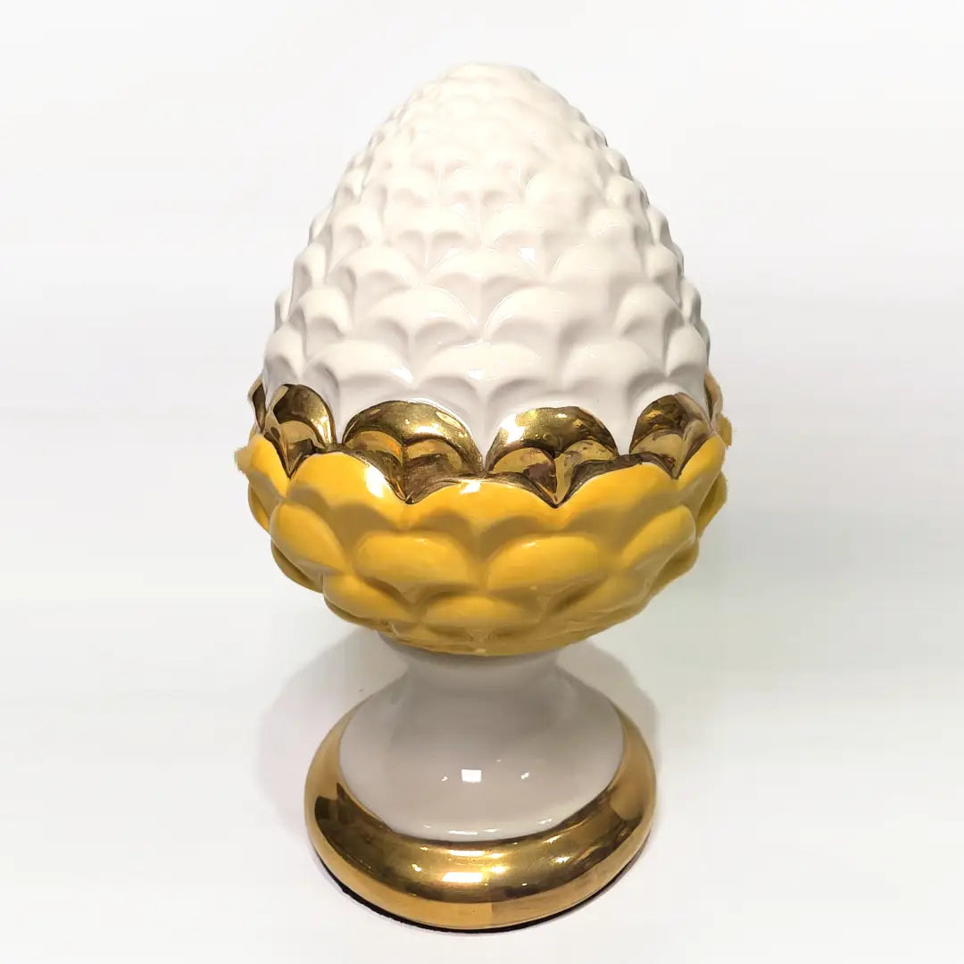 Pigna portafortuna in ceramica bicolore gialla e bianca lucida, impreziosita da una raffinata corona di squame dorate che ne definisce il profilo centrale. Questo elegante oggetto ornamentale artigianale poggia su una base circolare con finiture in oro, rappresentando un complemento d'arredo perfetto per decorazioni d'interni in stile chic e contemporaneo.
